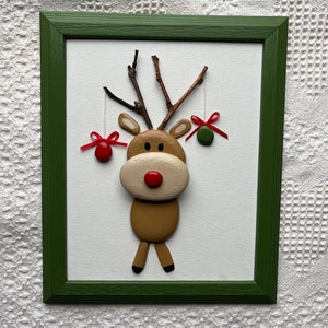 May include: A framed Christmas decoration depicting a reindeer crafted from natural materials. The reindeer has a brown body, beige face, and a red nose. Twigs form the antlers, and red and green baubles hang from them. The frame is green.