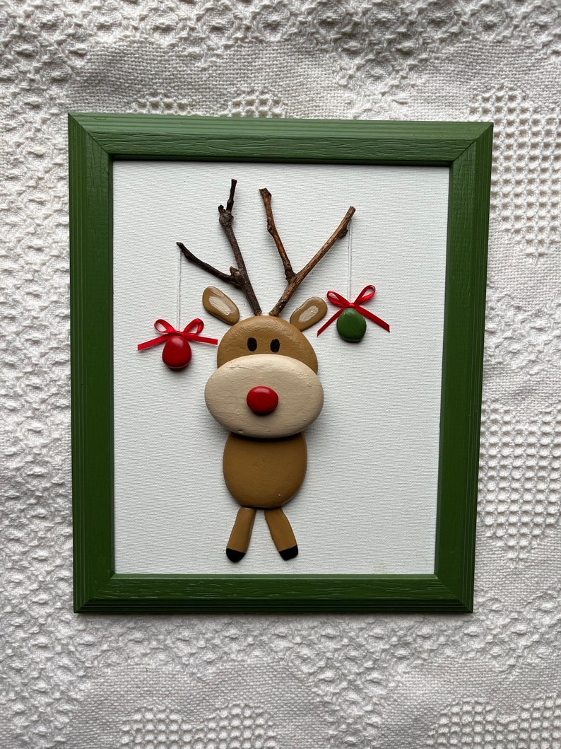 May include: A framed Christmas decoration depicting a reindeer crafted from natural materials. The reindeer has a brown body, beige face, and a red nose. Twigs form the antlers, and red and green baubles hang from them. The frame is green.
