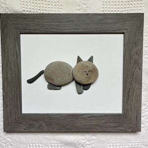 May include: A framed artwork featuring a cat made of smooth, gray stones. The cat has a round body and head, with a smiling face and pointed ears. The artwork is set against a white background and framed in a gray wooden frame.