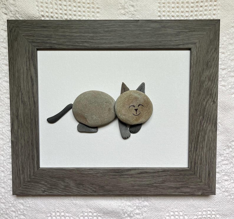May include: A framed artwork featuring a cat made of smooth, gray stones. The cat has a round body and head, with a smiling face and pointed ears. The artwork is set against a white background and framed in a gray wooden frame.