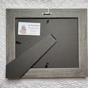 May include: Back of a gray framed picture with a dark gray backing and a kickstand. The frame has a textured wood grain appearance. A small white label with text and a stacked stone graphic is visible. A metal hanger is attached.