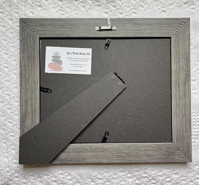 May include: Back of a gray framed picture with a dark gray backing and a kickstand. The frame has a textured wood grain appearance. A small white label with text and a stacked stone graphic is visible. A metal hanger is attached.