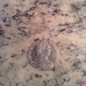 May include: A silver coin featuring a profile of a woman with a detailed hairstyle and inscription. The coin is circular and appears to be an antique or historical piece, resting on a speckled surface.