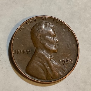 May include: A copper-colored 1956 US penny coin with the words "In God We Trust" and "Liberty" inscribed around the edge. The coin features a profile of Abraham Lincoln on the obverse side.