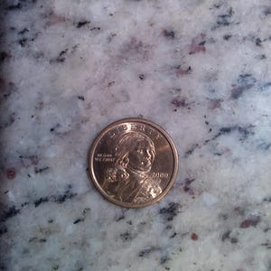 May include: A golden-colored Sacagawea dollar coin from the year 2000. The coin features the word "LIBERTY" at the top and the inscription "IN GOD WE TRUST" on the left side. The coin has a portrait of Sacagawea.