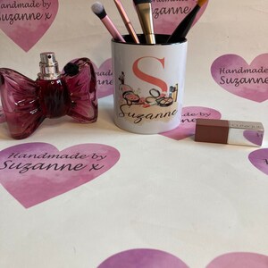 May include: A white ceramic mug with a black rim and a pink and orange design of makeup brushes, lipstick, and a compact. The mug has the letter "S" and the name "Suzanne" written on it. The mug is sitting on a white surface with a pink heart and the text "Handmade by Suzanne x" on it.