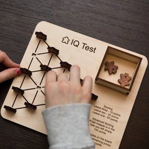 May include: A wooden IQ test game with a light wood board, black lines, and wooden game pieces. The board has the text "IQ Test" and scoring instructions. A person's hands are interacting with the game.