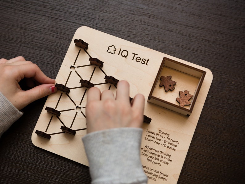 May include: A wooden IQ test game with a light wood board, black lines, and wooden game pieces. The board has the text "IQ Test" and scoring instructions. A person's hands are interacting with the game.