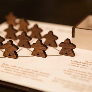 May include: A wooden board game with dark brown player pieces arranged on a light-colored wooden board. The game pieces are shaped like stylized people. The board has etched lines and text, including scoring instructions. A wooden box holds extra pieces.