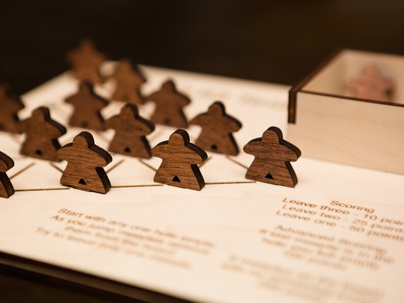 May include: A wooden board game with dark brown player pieces arranged on a light-colored wooden board. The game pieces are shaped like stylized people. The board has etched lines and text, including scoring instructions. A wooden box holds extra pieces.