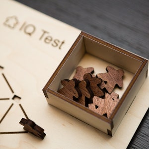 May include: Wooden IQ test game with a light wood base and a dark wood box containing several small wooden figures. The words "IQ Test" are etched into the base. A single figure sits outside the box.