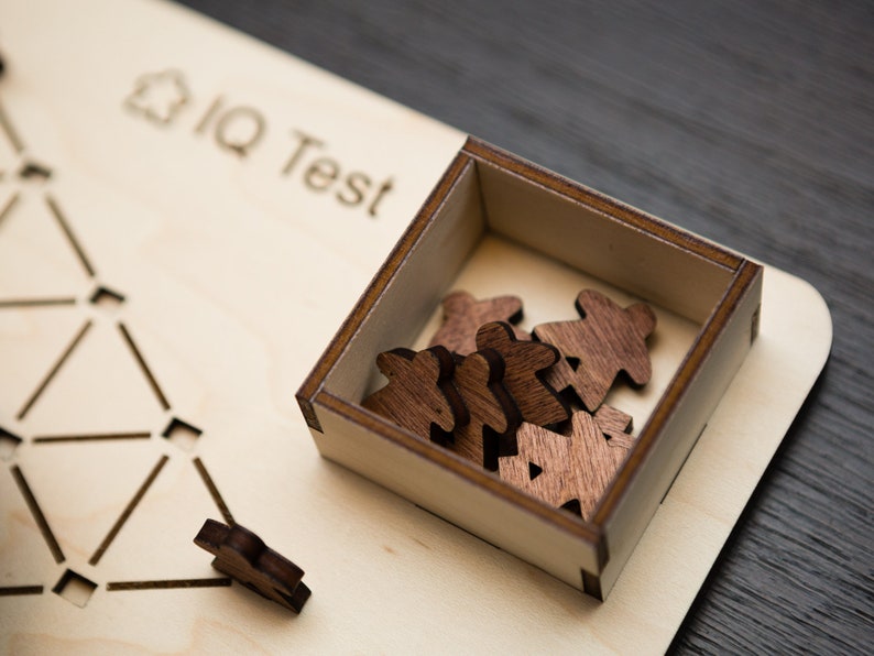 May include: Wooden IQ test game with a light wood base and a dark wood box containing several small wooden figures. The words "IQ Test" are etched into the base. A single figure sits outside the box.