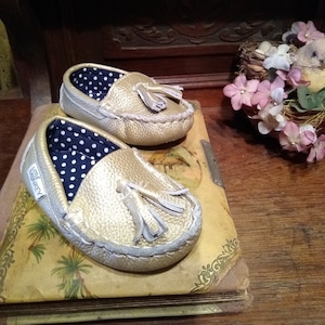 May include: A pair of gold leather moccasins with tassels. The moccasins have a black and white polka dot lining. The moccasins are sitting on top of an old book.