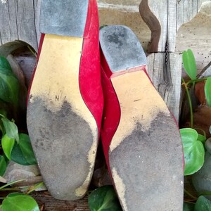 May include: Pair of red leather shoes with square toes, showing the soles. The soles are worn and dirty, with a light tan colour and a dark grey heel. The shoes are placed against a rustic wooden background with green foliage.