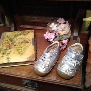 May include: Silver baby shoes with star detailing and Velcro straps are displayed on a wooden surface. A vintage photo album with floral designs and a decorative bird's nest with flowers are also visible. The scene is set on a wooden shelf.