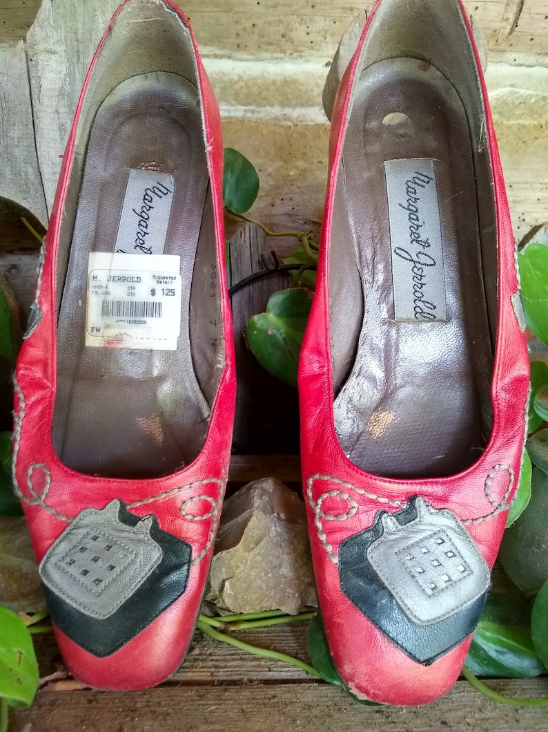 May include: Pair of red leather flats with decorative stitching and a unique design on the toe. The shoes have a label inside that reads "Margaret Jerrold". The flats are a size 7 and appear to be vintage.