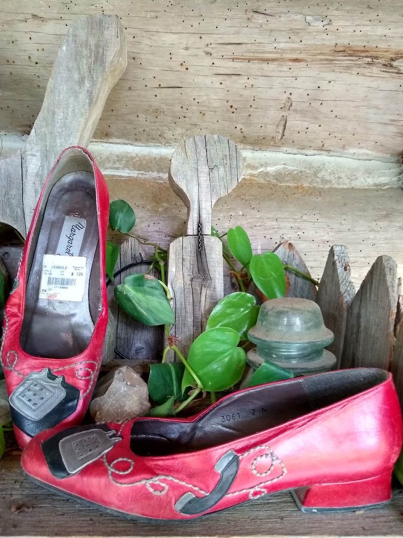 May include: Red leather flats featuring a telephone design. The shoes have a low heel and a black and grey telephone graphic on the toe. The shoes are displayed on a wooden surface.