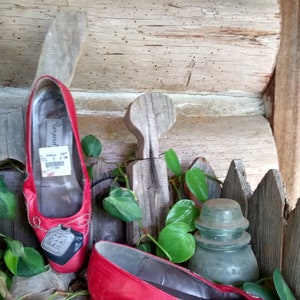 May include: A pair of vintage red leather pumps with a telephone design on the toe. The shoes are displayed on a rustic wooden surface with greenery and a glass insulator. The heels are approximately 3.8 cm.