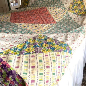 May include: A colorful patchwork quilt with a variety of floral and geometric patterns. The quilt features a mix of fabrics in shades of pink, blue, yellow, and purple. The quilt is spread over a bed with a white bed skirt.