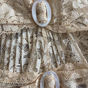 May include: Close-up of a cream-colored lace fabric with intricate floral patterns. Two oval-shaped, off-white buttons with a textured center are visible. The delicate lace and button details suggest a vintage or handmade garment.