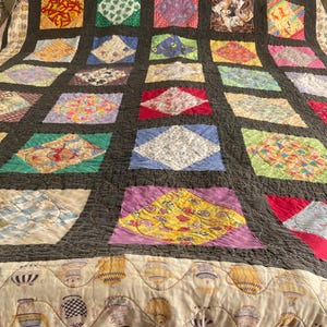 May include: A patchwork quilt with a dark gray background and a grid of colorful squares and diamond shapes. The squares feature various patterns and colors, including floral, geometric, and abstract designs. The quilt's border has a cream-colored fabric with a repeating pattern of hot air balloons.