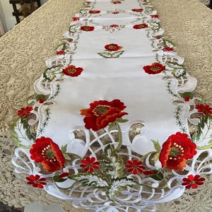 May include: White table runner with embroidered red poppies and green leaves. The runner features a scalloped edge with intricate cutwork and floral designs. A decorative piece for a dining table or sideboard.