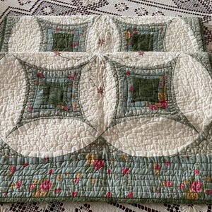 May include: A quilted fabric piece featuring a pattern of cream-colored circles and squares in shades of sage green, olive, and floral prints. The design includes intricate stitching, embroidered floral accents, and a bottom border with a rose pattern.