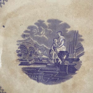 May include: A vintage blue and white ceramic plate with a detailed illustration. The central image depicts two figures in a classical setting, surrounded by architectural elements and a landscape. The plate's rim features a decorative border.