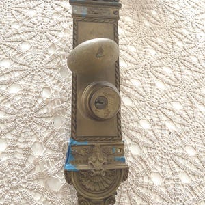 May include: Antique brass door handle set with an oval knob and keyhole. The rectangular backplate features ornate detailing, including a decorative top and bottom. The handle shows signs of age with a weathered appearance and blue paint.