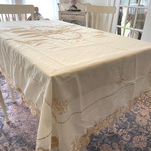 May include: Cream-colored tablecloth with embroidered floral details. The tablecloth is draped over a white table, showcasing intricate stitching along the edges and a central design. The floral embroidery is in a light tan color.