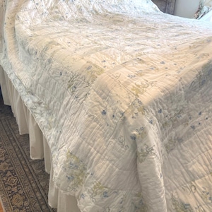 May include: A white quilted bedspread with a floral pattern in blue and green covers a bed. The bedspread has a ruffled edge and is draped over a bed skirt. The overall theme is a vintage, cottagecore aesthetic.