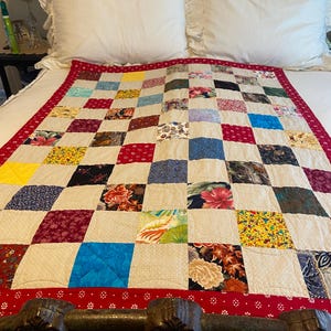 May include: A handmade patchwork quilt with a red border and a variety of colorful fabric squares. The quilt features floral, geometric, and solid patterns in shades of red, blue, yellow, and green. The quilt is displayed on a white bed.