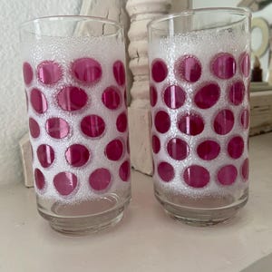 May include: Two clear glass drinking glasses with a pink polka dot pattern. The glasses have a white background with pink circles.