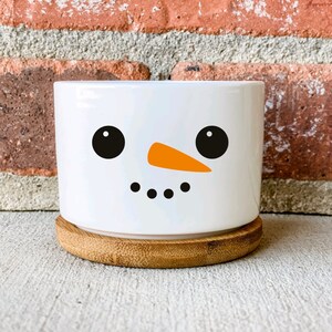 May include: White ceramic planter with a snowman face design. The snowman has black eyes, an orange carrot nose, and three black buttons. The planter sits on a wooden base.