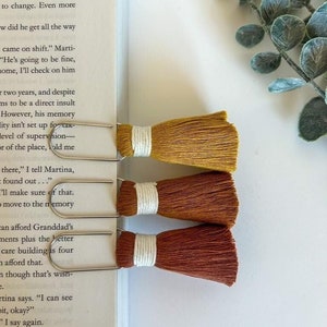 May include: Three metal bookmark clips with colorful tassels. The tassels are mustard yellow, burnt orange, and rust red. The tassels are made of yarn and have a white cord at the top.