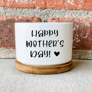 Puede incluir: Maceta de cer&aacute;mica blanca con base de bamb&uacute;. La maceta tiene el texto "Happy Mother's Day!" con un peque&ntilde;o coraz&oacute;n negro.