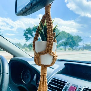 May include: A small potted plant hanging from the rearview mirror of a car. The plant is in a white pot and is surrounded by a macrame plant hanger made of brown cord. The plant hanger has a tassel at the bottom.