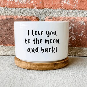 Puede incluir: Maceta de cer&aacute;mica blanca con base de bamb&uacute;. La maceta tiene el texto "I love you to the moon and back!" impreso en ella.