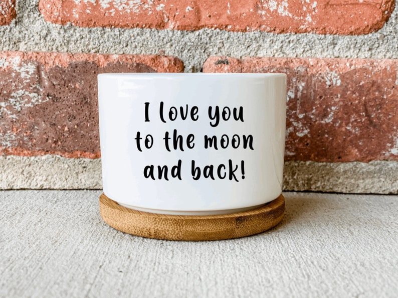 Puede incluir: Maceta de cer&aacute;mica blanca con base de bamb&uacute;. La maceta tiene el texto "I love you to the moon and back!" impreso en ella.