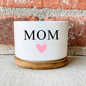 Puede incluir: Maceta de cer&aacute;mica blanca con la palabra "MOM" y un coraz&oacute;n rosa en ella. La maceta tiene una base de madera.