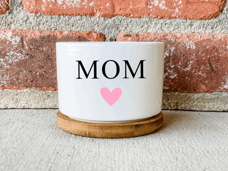 Puede incluir: Maceta de cer&aacute;mica blanca con la palabra "MOM" y un coraz&oacute;n rosa en ella. La maceta tiene una base de madera.