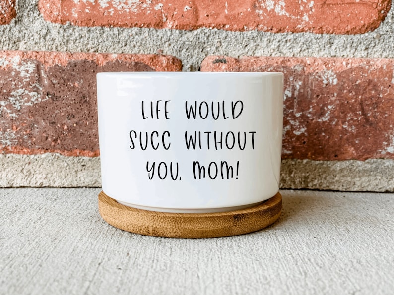 Puede incluir: Maceta de cer&aacute;mica blanca con base de madera. La maceta tiene el texto "Life would succ without you, Mom!" impreso en ella.
