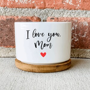 Puede incluir: Maceta de cer&aacute;mica blanca con base de bamb&uacute;. La maceta tiene el texto "I love you, Mom" en letras negras y un peque&ntilde;o coraz&oacute;n rojo.