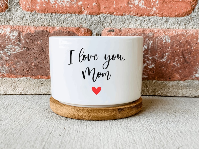 Puede incluir: Maceta de cer&aacute;mica blanca con base de bamb&uacute;. La maceta tiene el texto "I love you, Mom" en letras negras y un peque&ntilde;o coraz&oacute;n rojo.