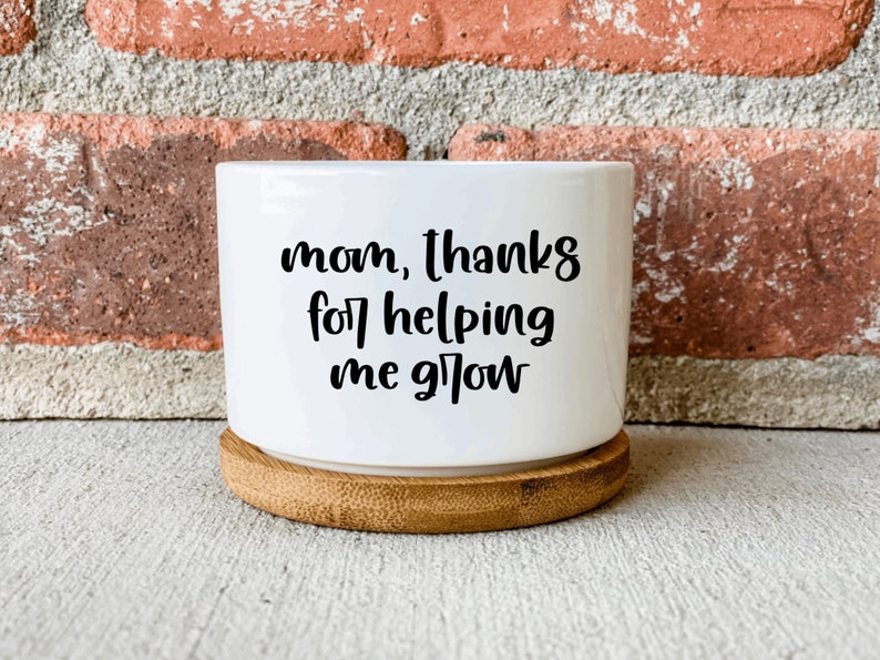 Puede incluir: Maceta de cer&aacute;mica blanca con base de madera. La maceta tiene texto negro que dice "mam&aacute;, gracias por ayudarme a crecer".