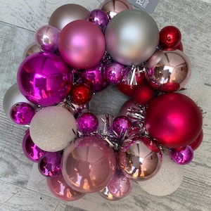 May include: A festive Christmas wreath composed of various sized ornaments in shades of pink, silver, and red. The wreath includes shiny and matte spheres, with a silver ribbon for hanging. The tag reads "RAZ Imports".