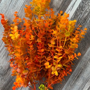 May include: A bundle of artificial eucalyptus stems in shades of orange, yellow, and green. The leaves are small and rounded, with a textured appearance. The stems are arranged to create a full, bushy look, perfect for fall decor.