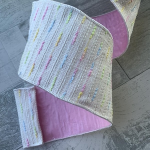 May include: White ribbon with a pink backing and a colorful confetti pattern. The ribbon is woven with small, colorful dots.