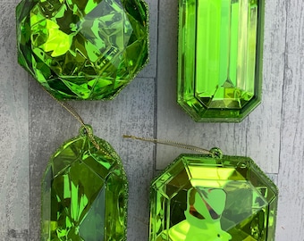 Puede incluir: Cuatro adornos navideños verdes facetados en varias formas: una esfera, un rectángulo, un óvalo y un octágono. Cada adorno tiene una cuerda dorada para colgar y un borde verde brillante. Dispuestos sobre una superficie de vetas de madera gris claro y blanco.