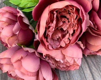 May include: A bouquet of artificial pink peonies with green leaves. The peonies are arranged in a cluster, with the blooms facing outward. The flowers are a soft, dusty pink color, and the leaves are a vibrant green.